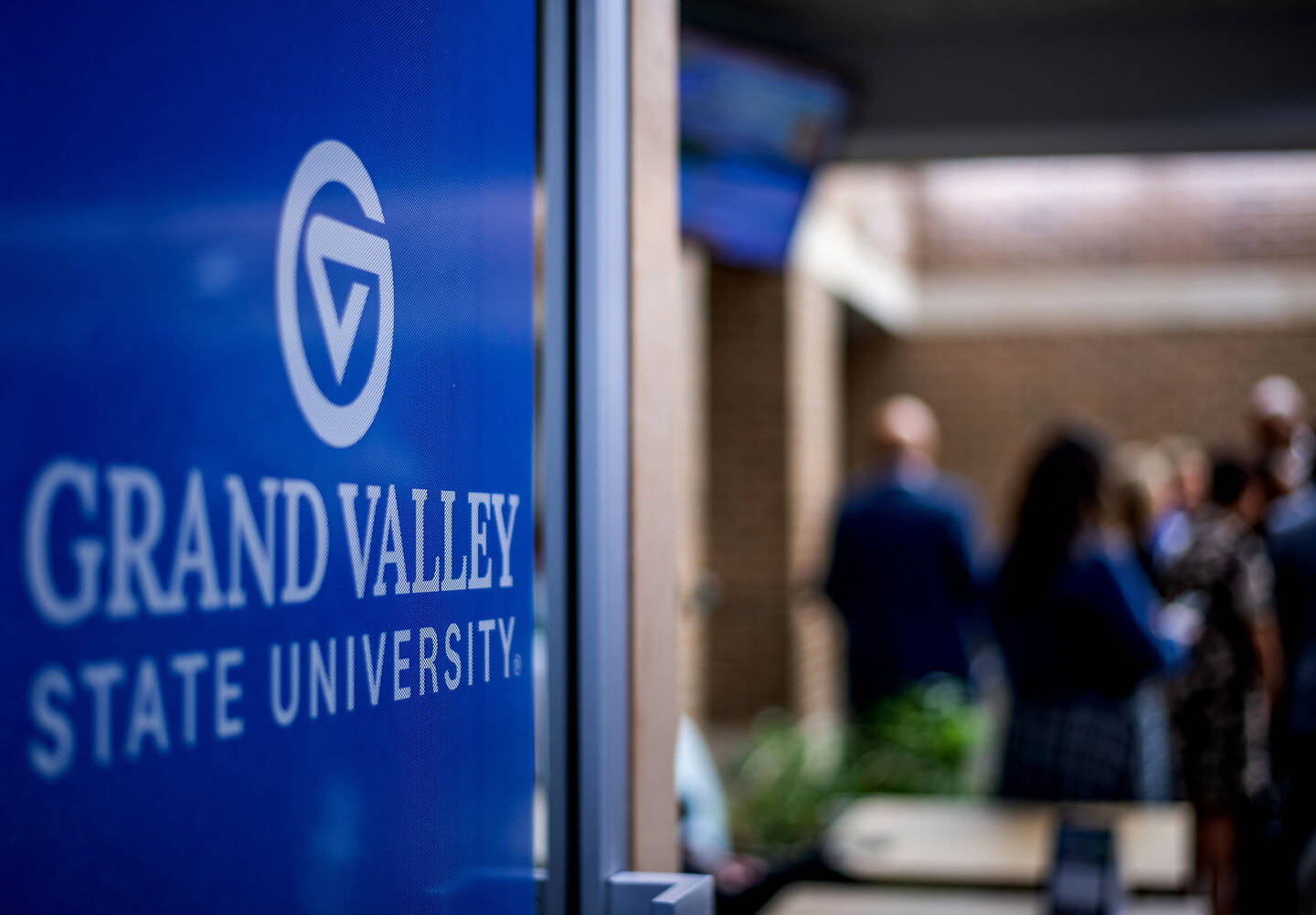 A blue sign for Grand Valley State University is in focus, with blurred figures of people in the background.